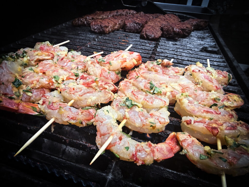 Denver steaks searing on a charcoal grill with shrimp skewers on the cooler side of a two-zone fire