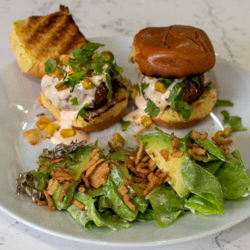 Elote smash sliders topped with charred corn and lime sauce on a cast iron skillet on a Weber grill